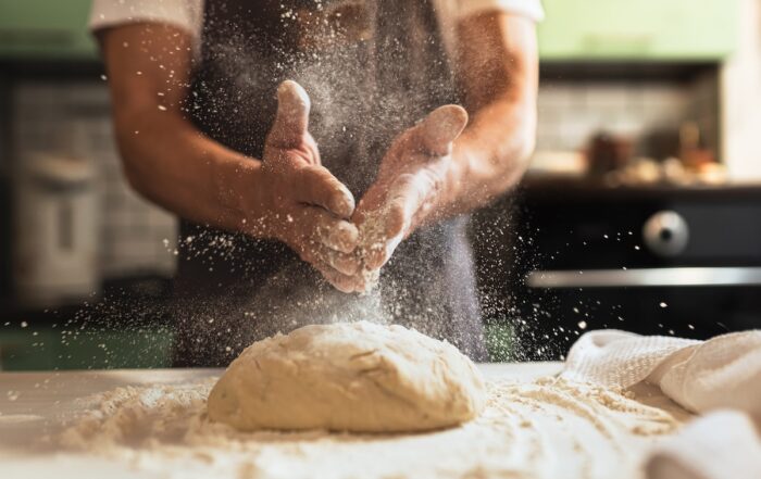 hands sprinkling flour over dough on bakery bench