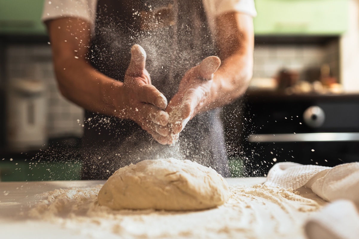 hands sprinkling flour over dough on bakery bench